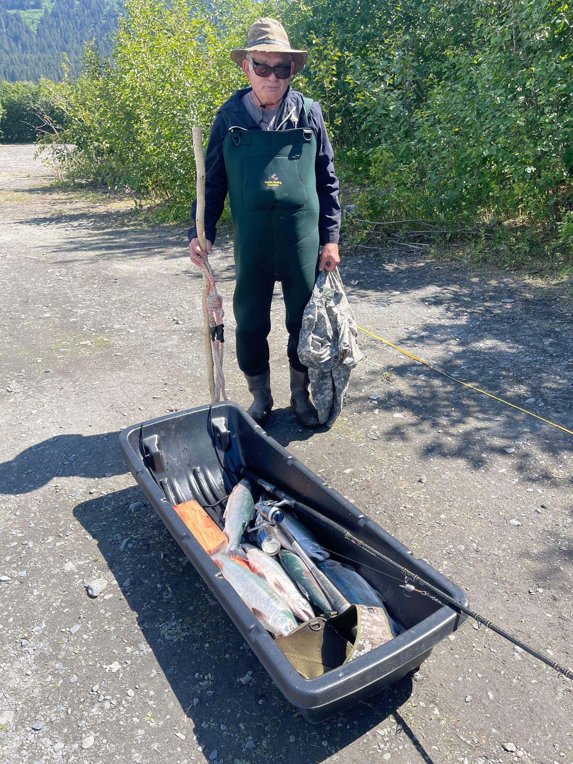 A person in chest waders and a hat stands on a gravel path, holding a fishing rod beside a sled filled with fresh fish.