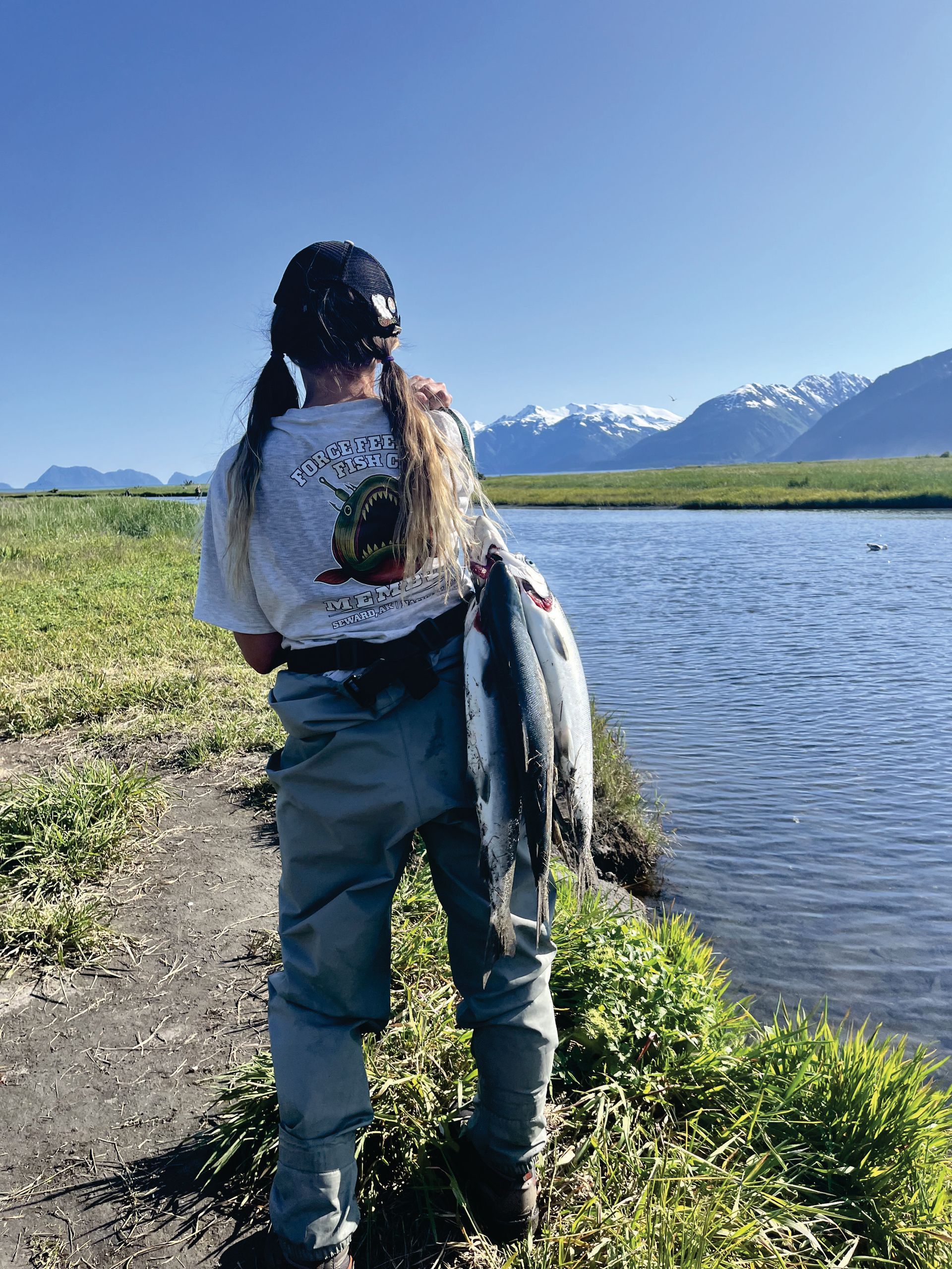 Person holding two fish by a stream, mountains in background.