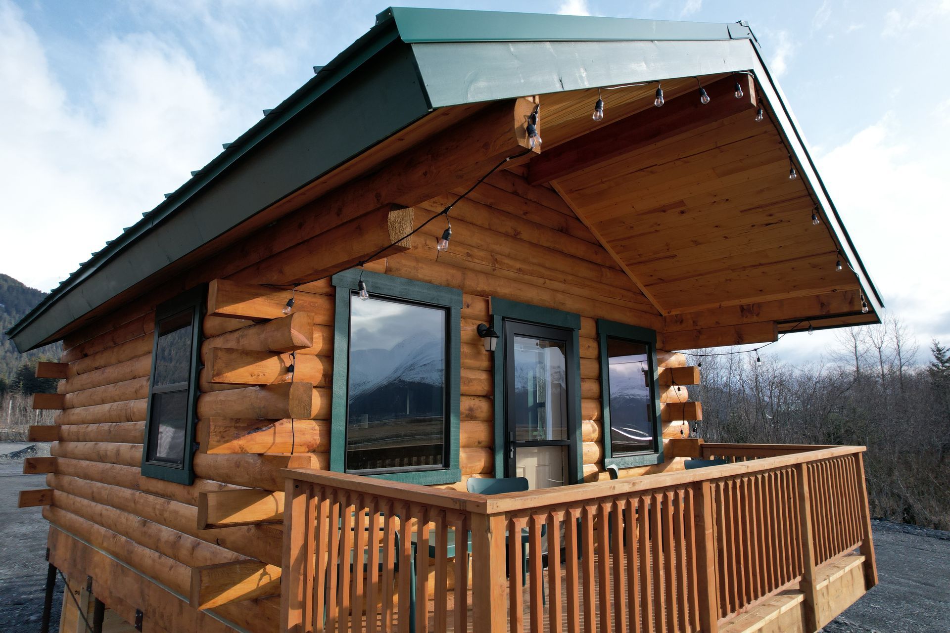 Log cabin with a wooden deck, green trim, and string lights on a sunny day.