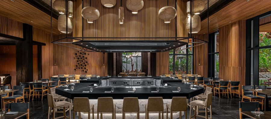 Un interior de restaurante japonés moderno con detalles de madera, una isla de cocina central y luces colgantes.