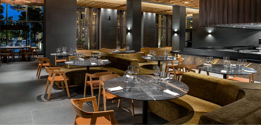 Interior de restaurante con mesas redondas dispuestas para cenar, sillas de madera y una cabina larga y curva.