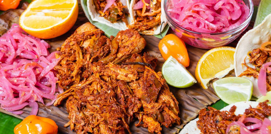 Una colorida variedad de comida mexicana, que incluye tacos, cebollas encurtidas, frutas y carne desmenuzada.