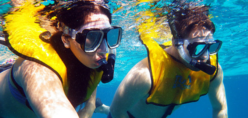 Dos personas haciendo snorkel en aguas azules, usando máscaras, tubos y chalecos salvavidas amarillos.
