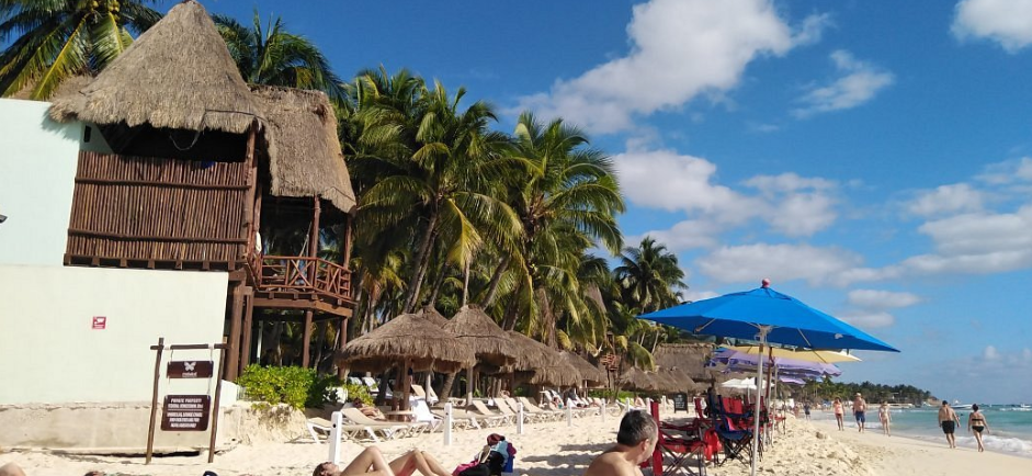 Escena de playa con estructuras con techo de paja, palmeras, sombrillas azules, gente y arena blanca bajo un cielo brillante.