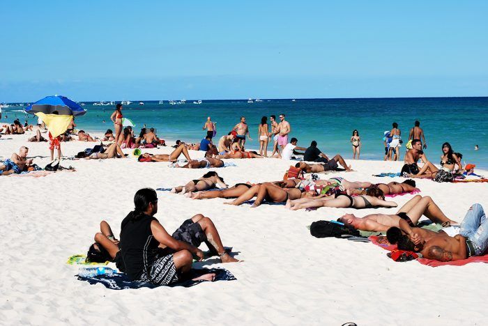 Gente tomando el sol y nadando en una concurrida playa de arena blanca con océano azul y cielo.