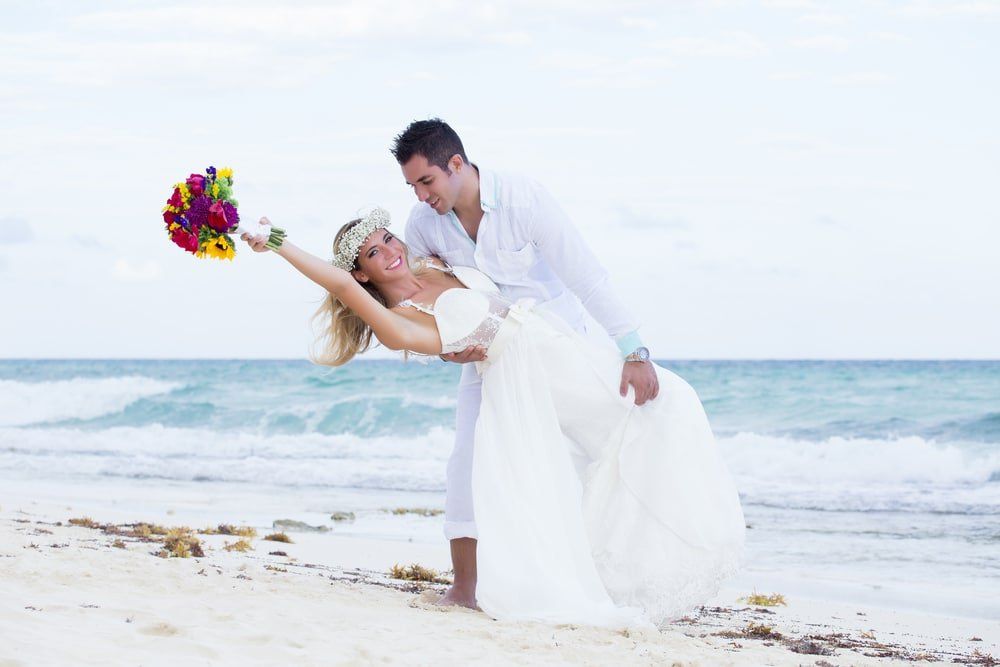 Couple dances on beach; bride in white dress, holding bouquet, leans back, groom in white shirt supports her.
