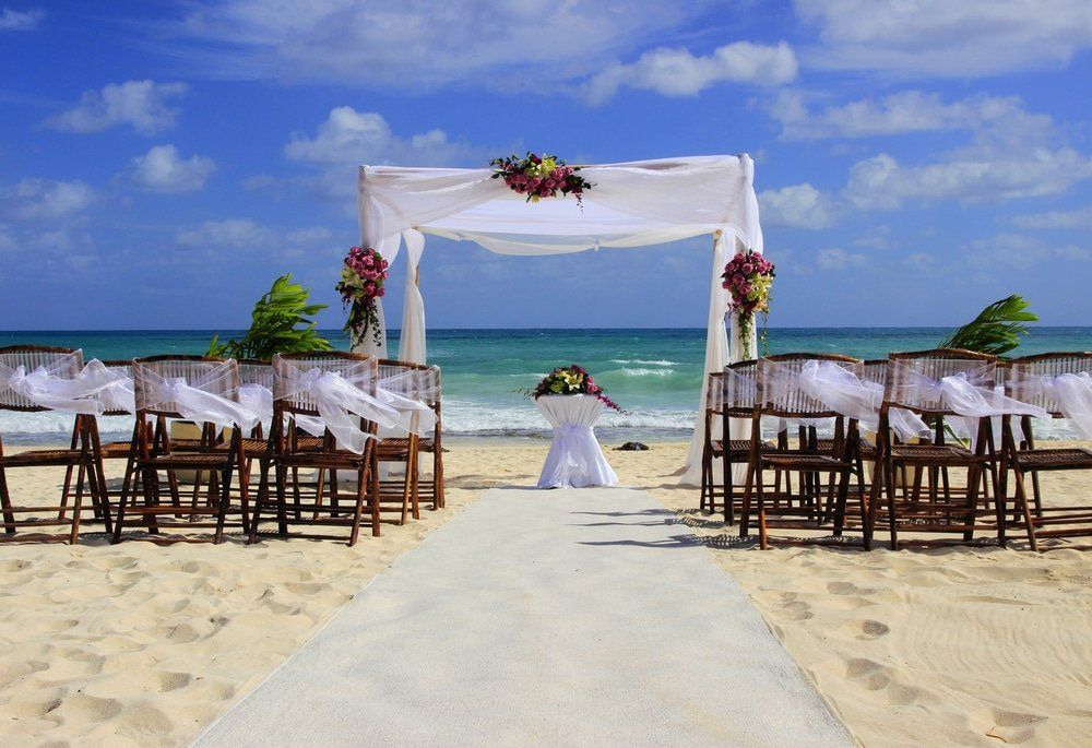 Beach wedding ceremony setup: arch with flowers, chairs, aisle on sand, ocean background.