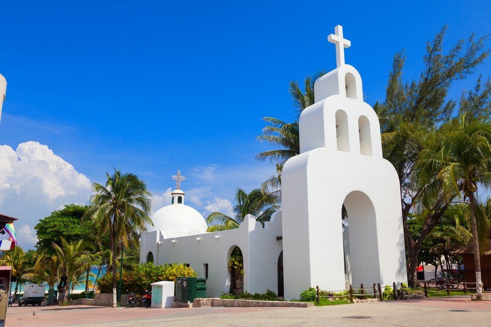White church with cross on top, under blue sky, near palm trees, possibly in a tropical location.