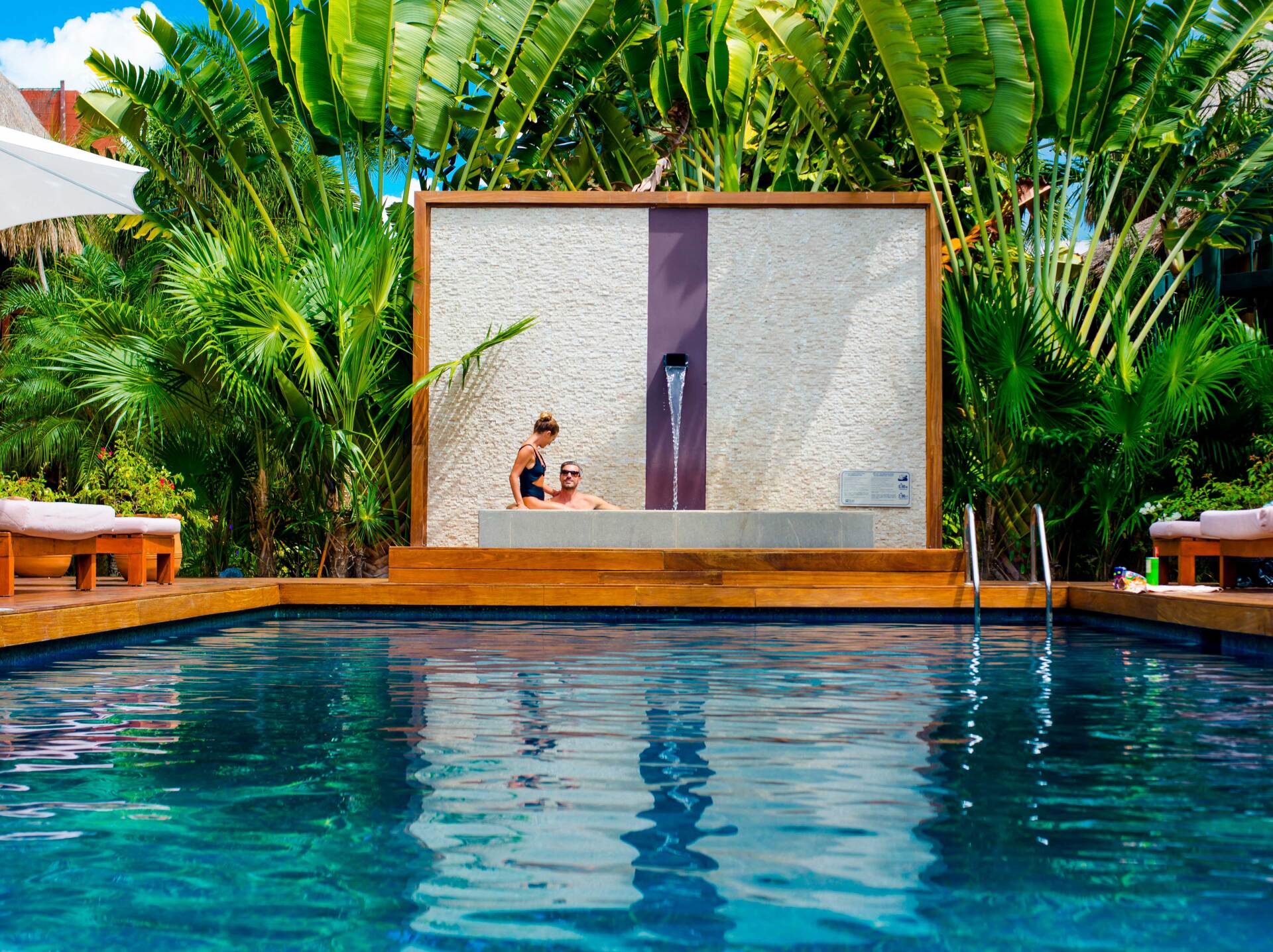 Pool with mosaic wall and two people. Lush tropical foliage surrounds a wooden deck with lounge chairs.