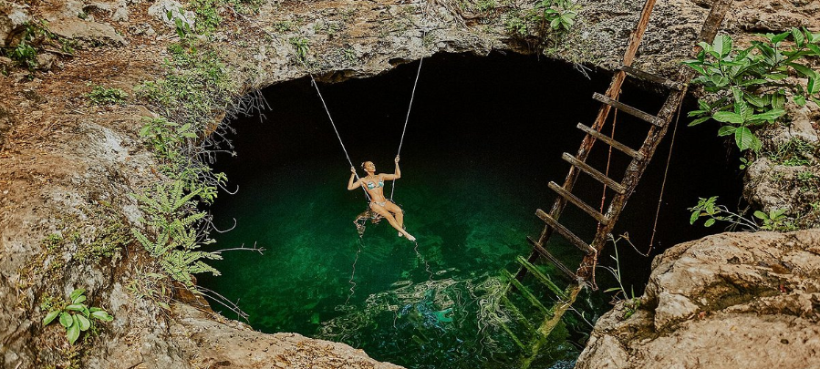 Persona se balancea en un cenote turquesa, junto a una escalera de madera y plantas verdes.