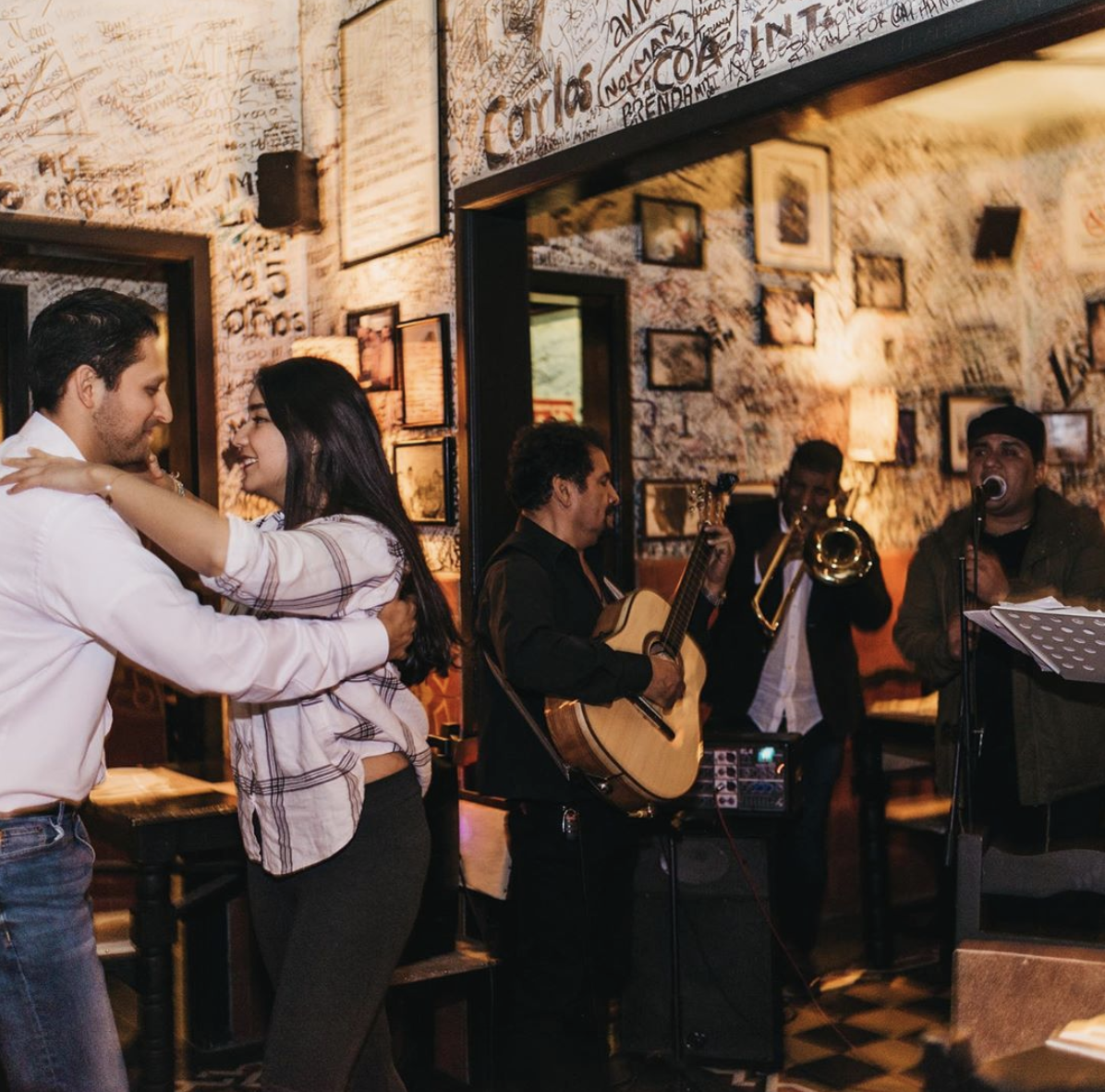 Una pareja baila al ritmo de música en vivo en un bar ; la banda toca la guitarra, la trompeta y canta.