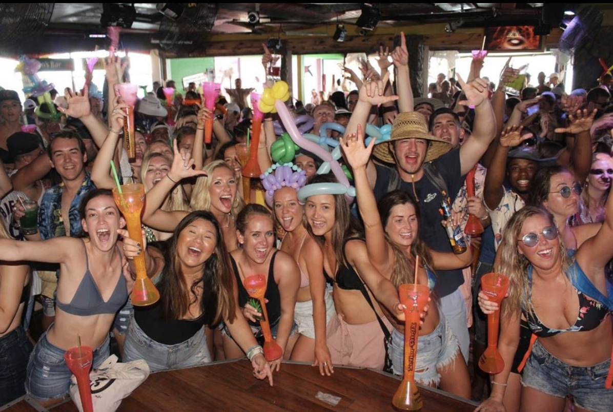 Grupo de personas de fiesta en un bar, con los brazos alzados y bebidas en mano. Colores brillantes, ambiente animado.