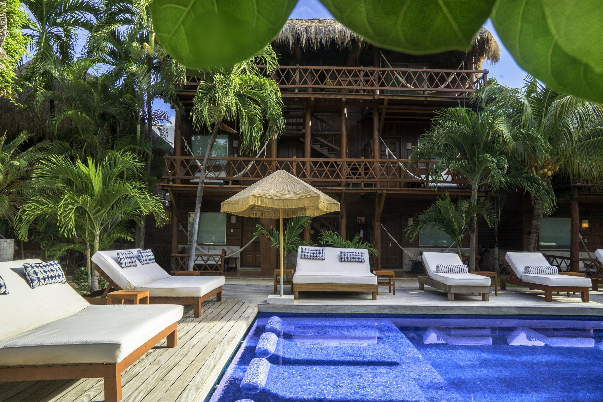 Poolside resort with wooden building, thatched roof, lounge chairs, and palm trees.