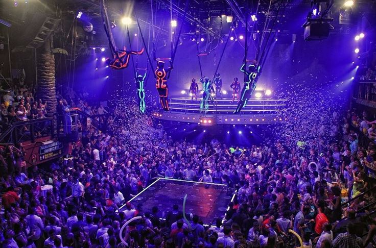 Gran discoteca abarrotada de gente, con bailarines en cables suspendidos sobre la pista de baile, bañados por luces .