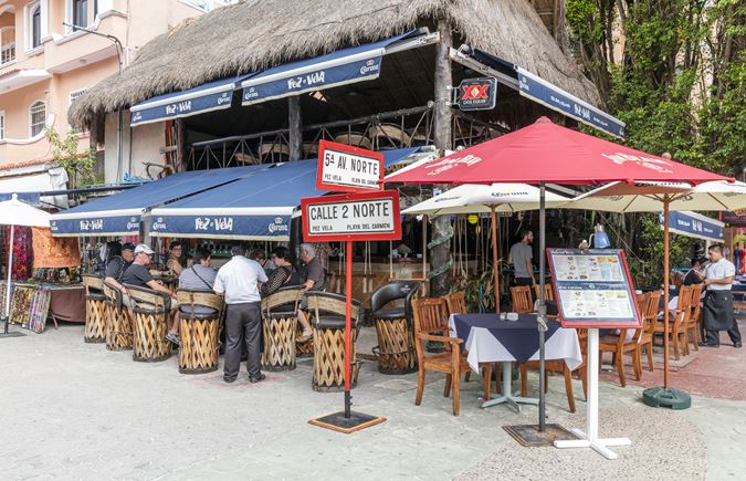 Restaurante con toldo azul, techo de paja, músicos tocando tambores, asientos al aire libre con sombrillas.