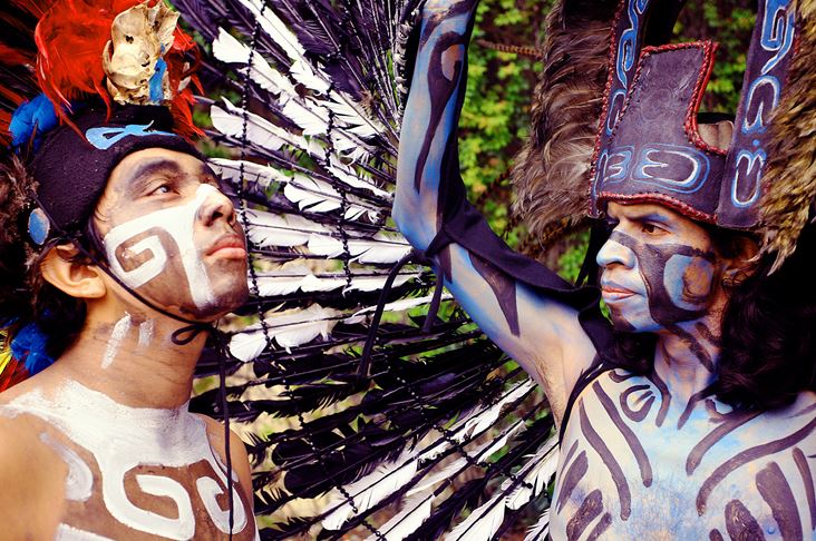 Dos personas con trajes ornamentados de estilo mesoamericano y pintura facial. Una con un tocado azul y blanco.