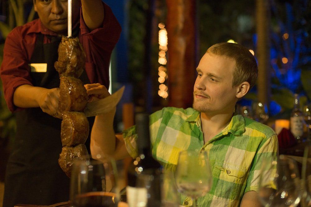 Man at table, watching server slice meat off skewer in outdoor restaurant.