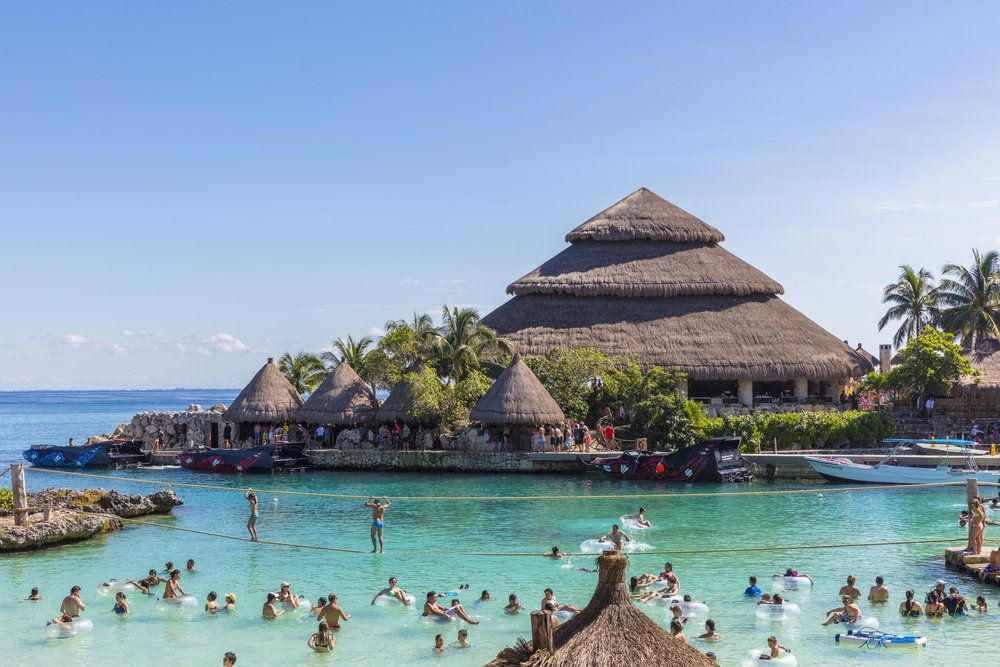 People swimming in turquoise water near thatched-roof buildings and palm trees on a sunny day.