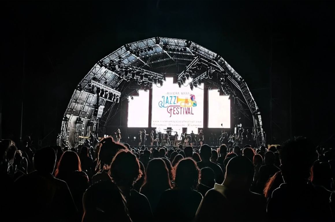 Escenario de conciertos por la noche con pantalla grande; multitud de personas recortadas en primer plano.