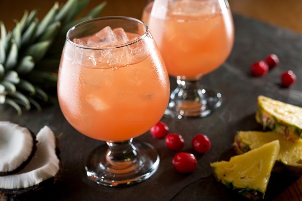 Two pink cocktails in stemmed glasses with ice, pineapple, coconut, and cranberries.