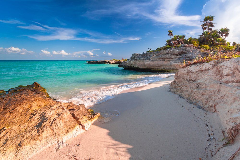 Beach with white sand, turquoise water, and rocky cliffs under a blue sky.