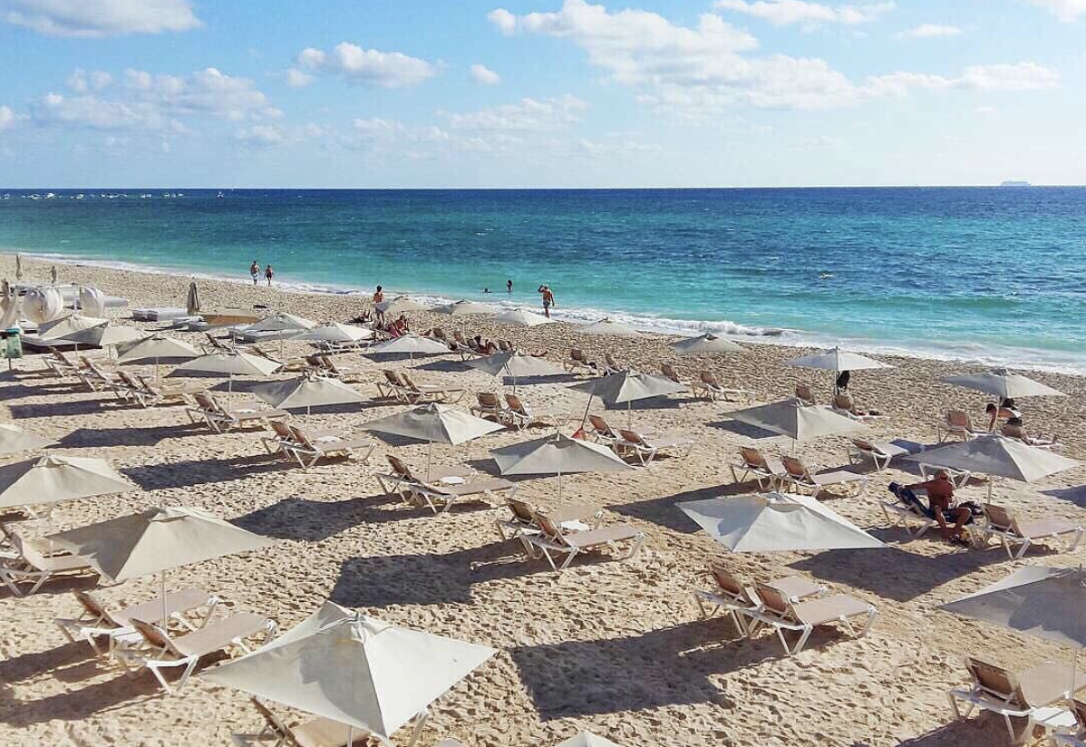 Beach with many lounge chairs and umbrellas, ocean in the background, blue sky with some clouds.