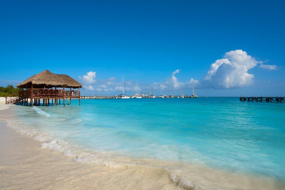 Beach scene with turquoise water, thatched roof structure, boats, and blue sky.