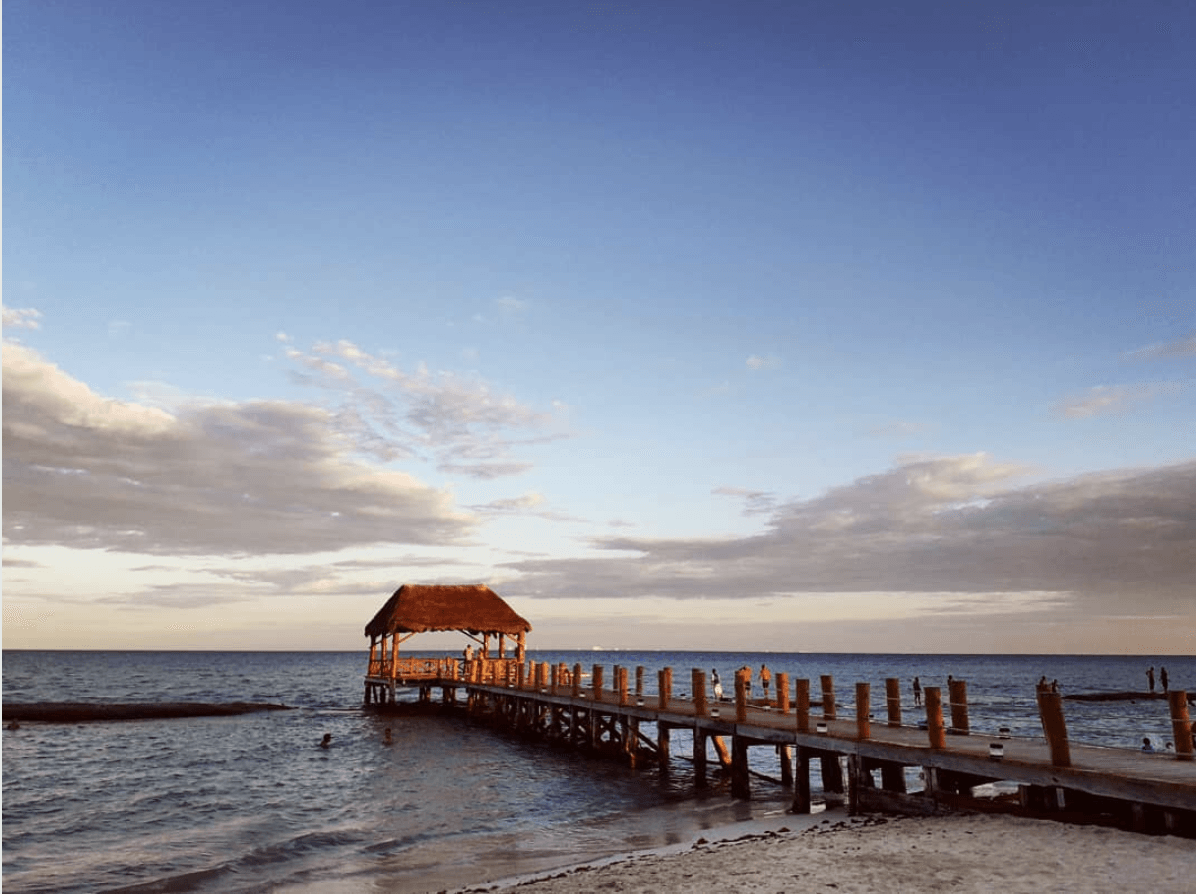 Wooden pier with thatched roof gazebo over calm ocean under a partly cloudy sky.