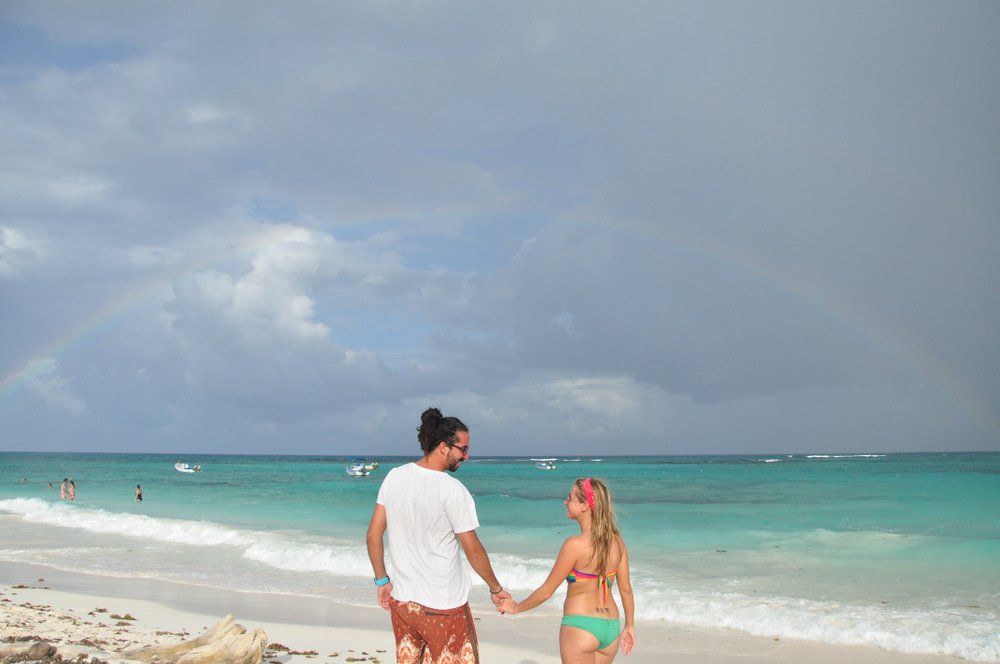Couple holding hands on beach; rainbow in the sky over turquoise water.