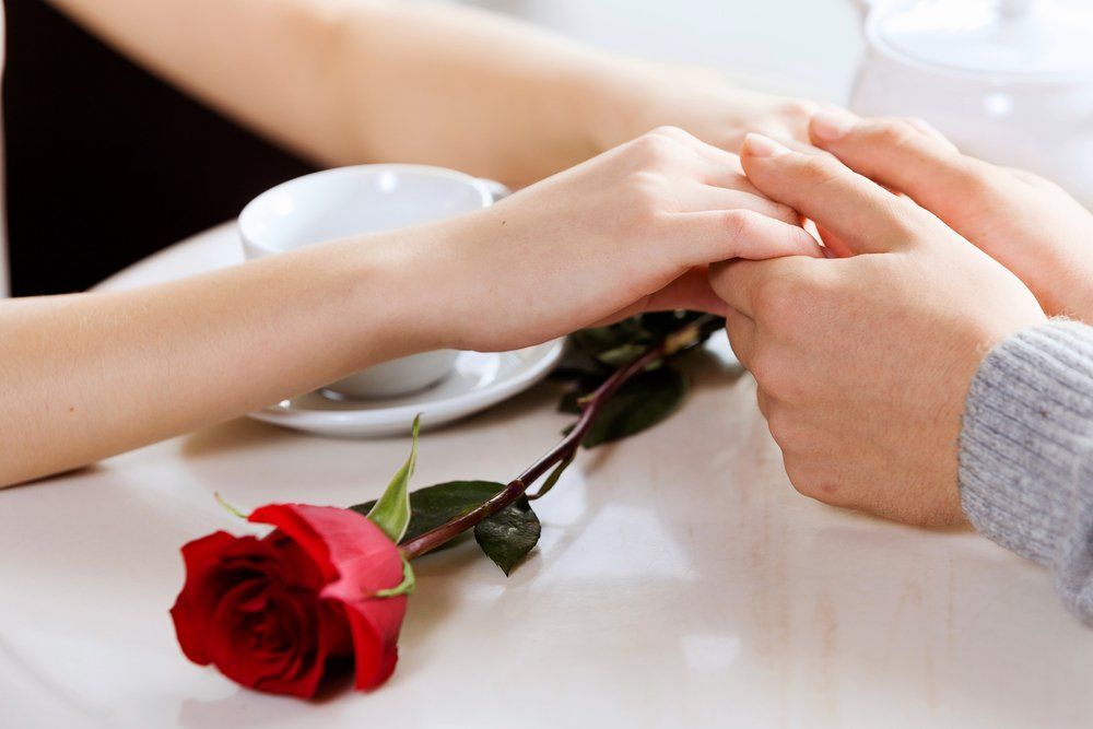 Hands clasped on a table; red rose and teacup nearby.