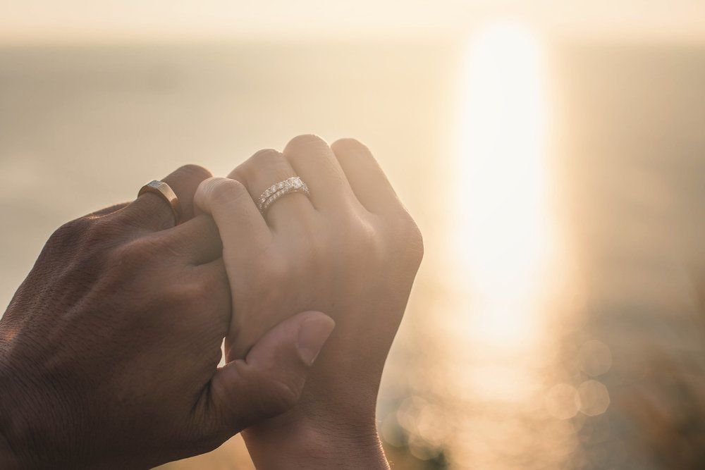 Hands clasped, wedding rings visible, with sunlight over water in background.