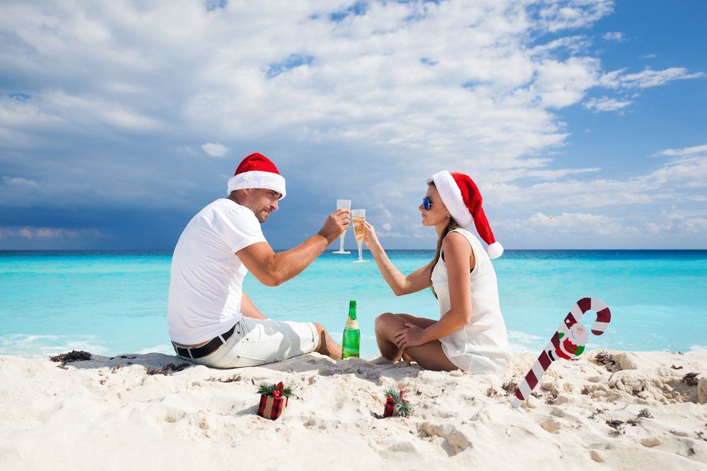 Pareja en la playa con gorros de Papá Noel brindando con champán. Escenario navideño, cielo azul y océano.