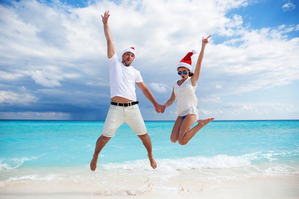 Couple wearing Santa hats, jumping for joy on a beach. Blue ocean and sky.