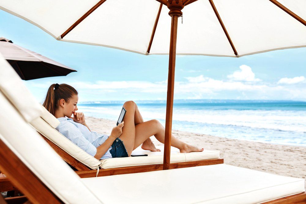 Woman relaxes on beach chair, reading a tablet under a white umbrella, ocean view.