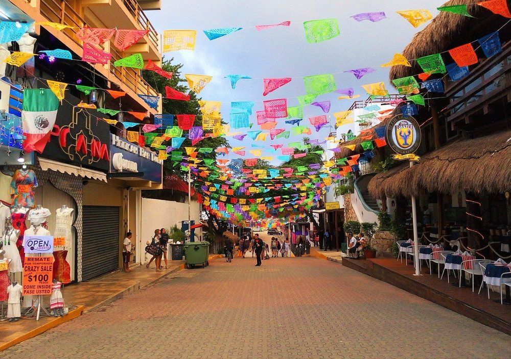 Street lined with shops, decorated with colorful papel picado flags. People walk on the cobblestone street.
