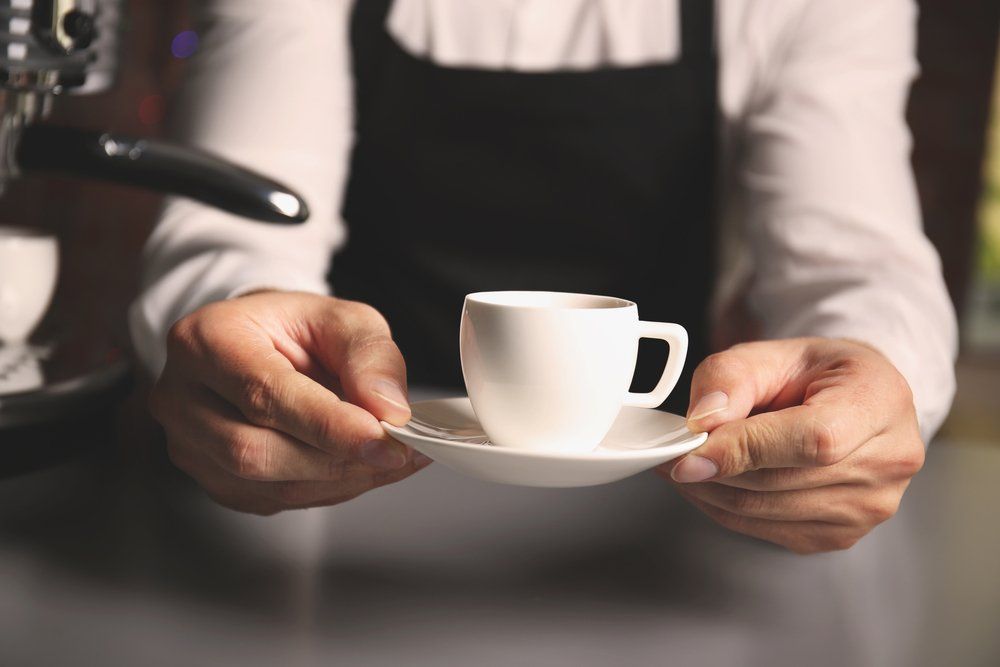 Barista holding a white coffee cup and saucer near an espresso machine.