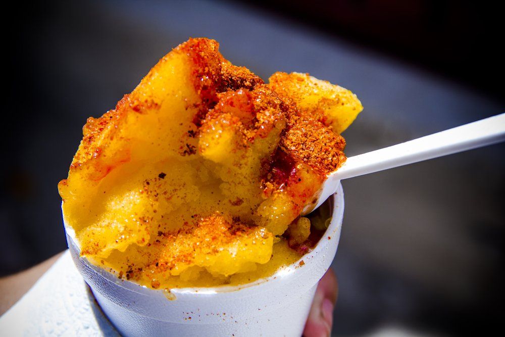 Yellow shaved ice in a white cup, topped with red chili powder, held with a white spoon.