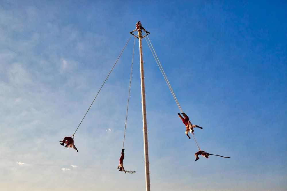 Voladores de Papantla ceremony: Men suspended from a tall pole, spinning downwards against a blue sky.