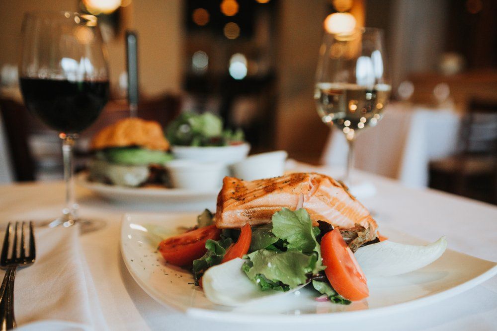 Plato de salmón con ensalada, copas de vino y hamburguesa sobre un mantel blanco en un entorno de restaurante.