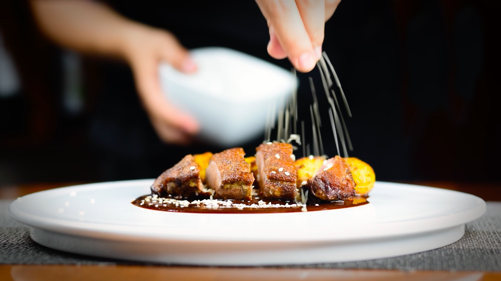 A chef sprinkles seasoning over sliced duck breast and golden potatoes on a white plate.