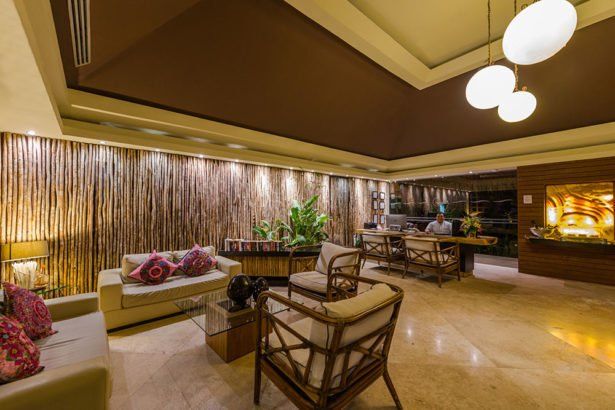 Lobby with cream sofa, rattan chairs, bamboo wall, plants, and desk with person at a wooden desk under brown ceiling.
