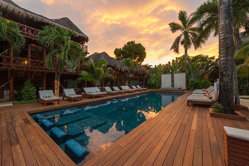 Swimming pool surrounded by wooden deck and lounge chairs, tropical resort at sunset.