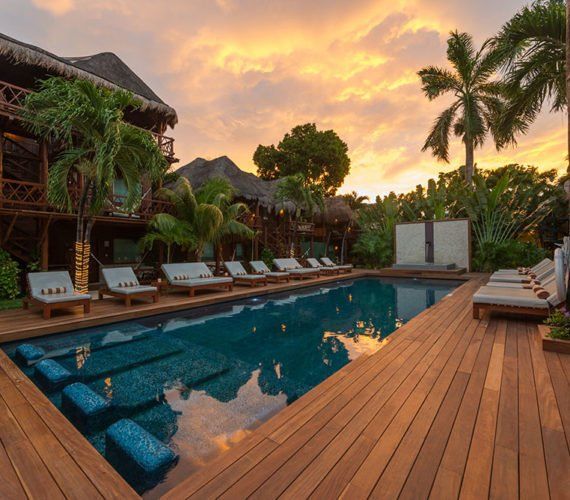Wooden deck surrounds a pool with blue tile steps. Palm trees and resort buildings under an orange sunset.