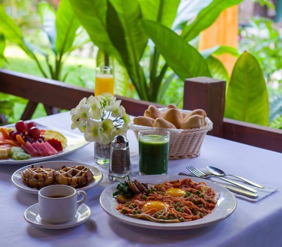 Breakfast spread on a white tablecloth, including fruit, pastries, eggs, juice, and flowers, outdoors with foliage.