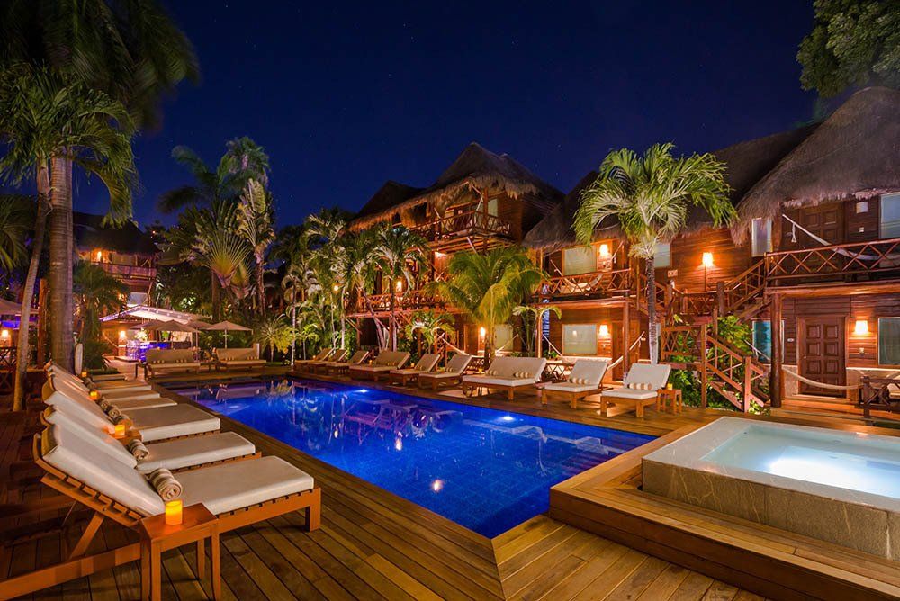 Night view of a resort pool area with lounge chairs and buildings with thatched roofs, palm trees, and lights.