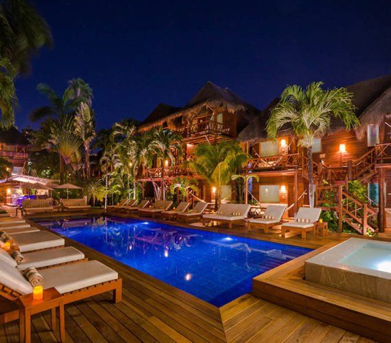 Night view of a luxury resort with pool, wooden deck, palm trees, and illuminated buildings.