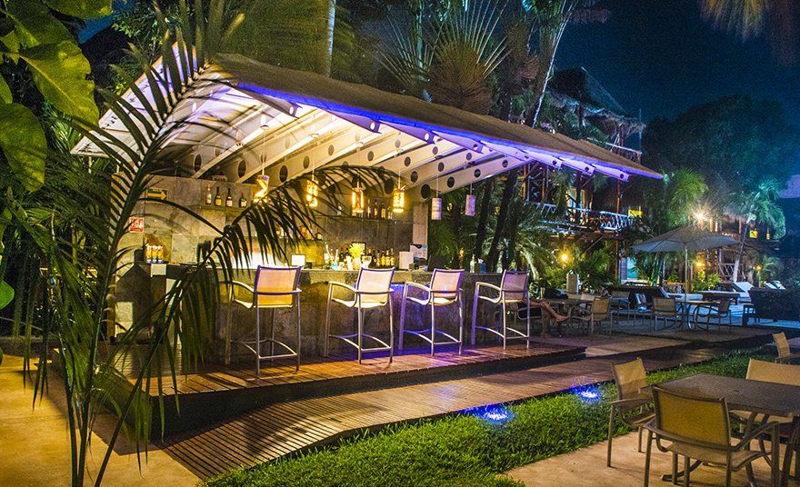 Outdoor bar at night with overhead lighting, stools, and tables in a tropical setting.