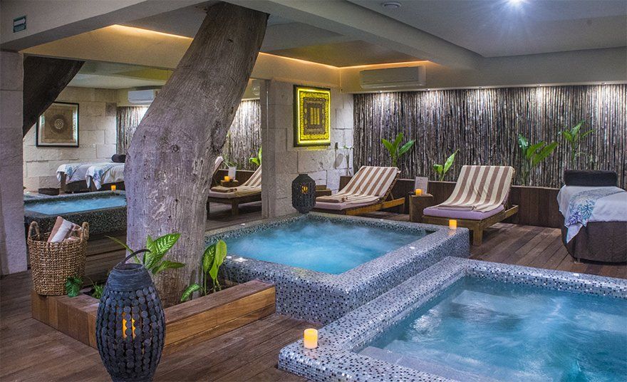 Spa with two hot tubs, lounge chairs, and massage tables, featuring a tree trunk and warm lighting.