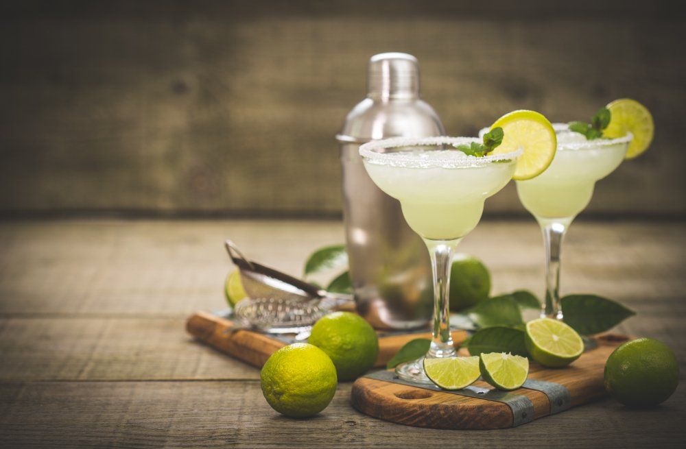 Dos margaritas con guarnición de lima, coctelera y limas sobre superficie de madera.