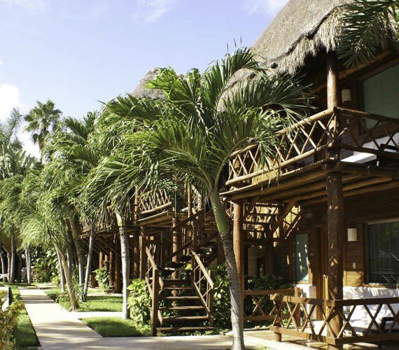 Row of tropical bungalows with wooden balconies and thatched roofs, palm trees, and walkway.